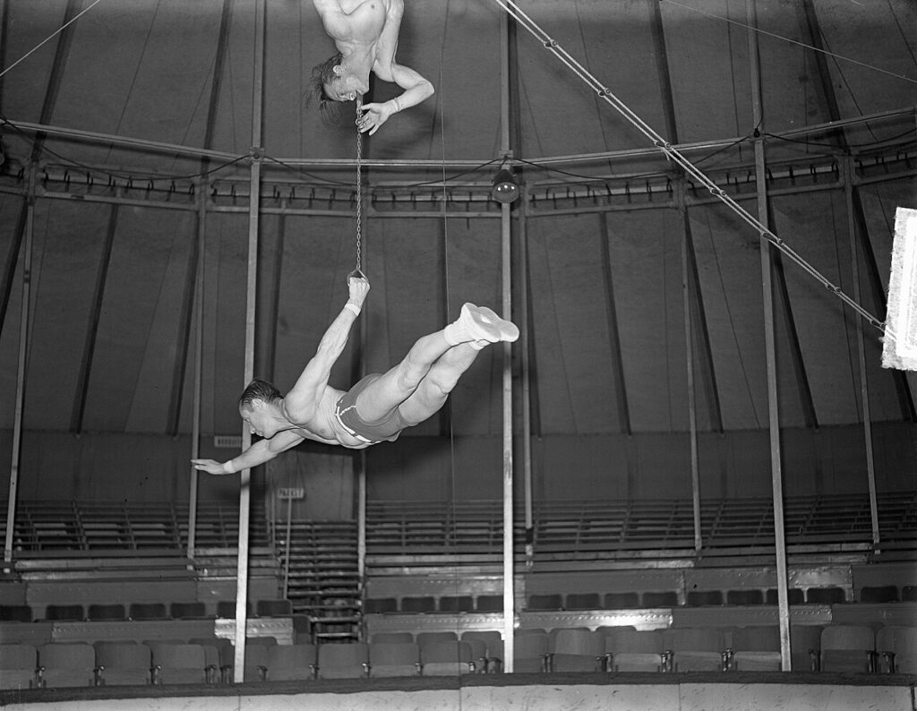 A man holding a chain in his mouth, another man dangling from the chain.