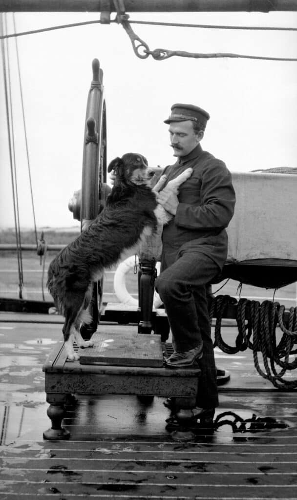 Dog posing standing up on his owner, both on a ship in front of the ship's steering wheel.